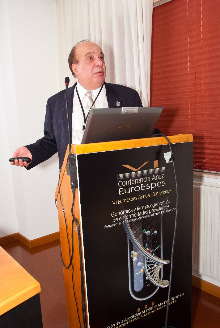 Una persona con traje habla mientras sostiene un dispositivo, de pie en un podio con un ordenador portátil y una pancarta de la VI Conferencia Anual de EuroEspes. La pancarta menciona la genómica y la farmacogenómica. Al fondo, unas persianas de madera están parcialmente abiertas.