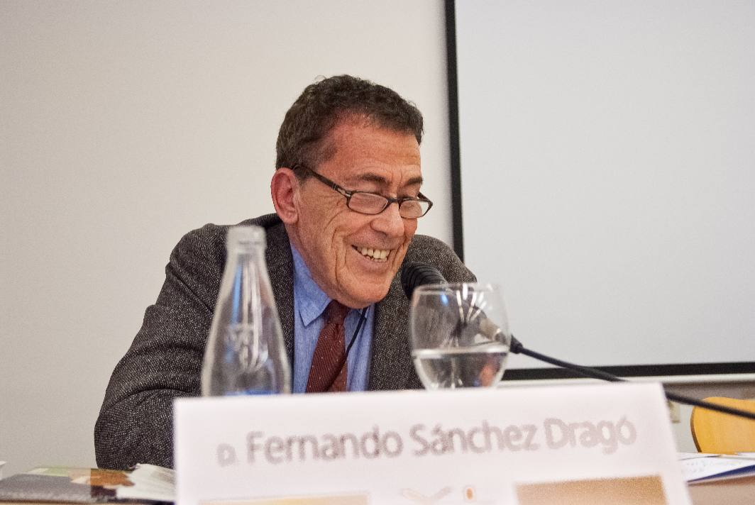 Un hombre está sentado a una mesa, sonriendo y hablando por un micrófono. Lleva gafas, una chaqueta gris y una camisa azul. Delante de él hay una placa con un nombre parcialmente visible, una botella de agua y un vaso de agua.