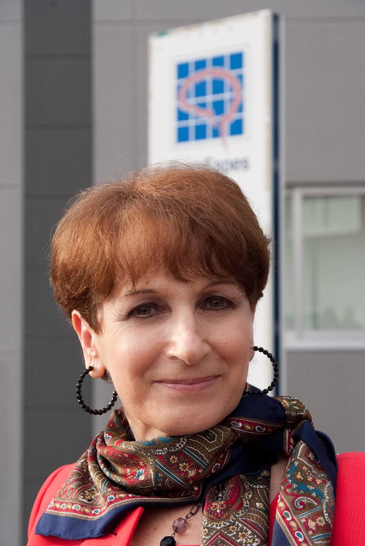 Una persona con cabello castaño corto y pendientes de aro sonríe. Lleva una bufanda estampada de colores y una camiseta roja. Al fondo hay un cartel borroso con un diseño azul y rosa.