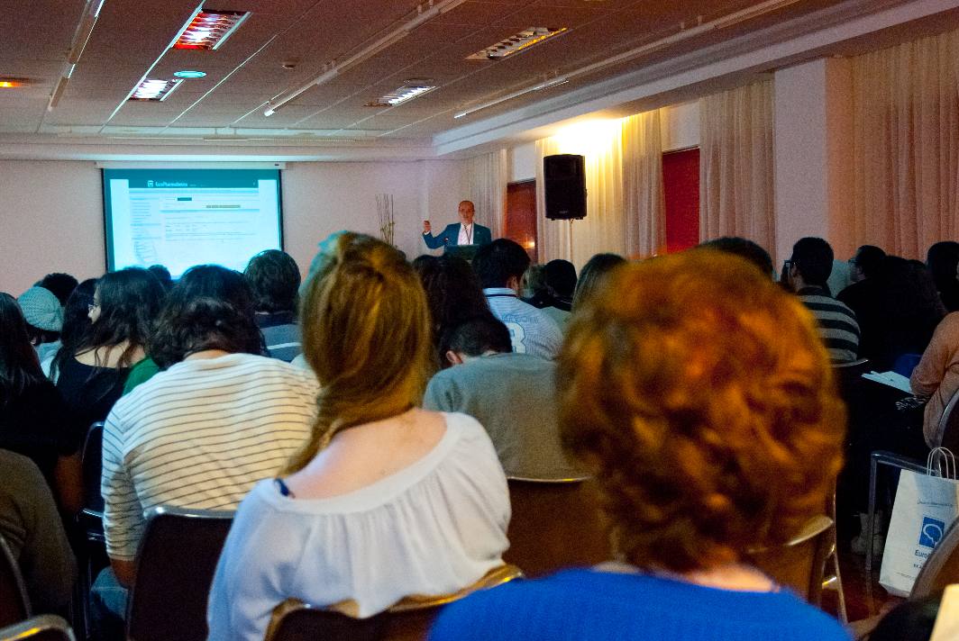 Público sentado en una sala, frente a un orador que se encuentra de pie junto a una presentación proyectada en una pantalla. La sala está suavemente iluminada y el orador hace gestos hacia la pantalla para interactuar con los asistentes.
