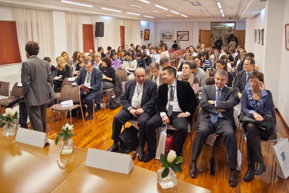 Un grupo de personas se sienta en una sala de conferencias, mirando hacia adelante. Algunas están conversando, mientras que otras miran documentos. La sala está muy iluminada y hay cuadros enmarcados en las paredes. Unas cuantas sillas vacías están cerca del frente.