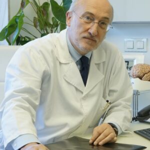 Un médico con bata blanca está sentado en un escritorio y mira a la cámara. Tiene la cabeza calva y gafas. Detrás de él hay plantas y un modelo de cerebro. Sostiene un documento o una película sobre la mesa.