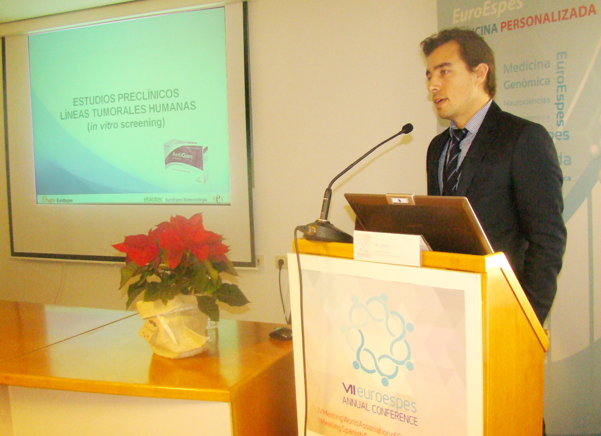 Un hombre con traje hace una presentación en un podio durante la VII Conferencia Anual de EuroEspes. En una pantalla se muestra una diapositiva titulada "Estudios preclínicos de línea tumoral humana (cribado in vitro)". Sobre la mesa hay una maceta con una flor de pascua.