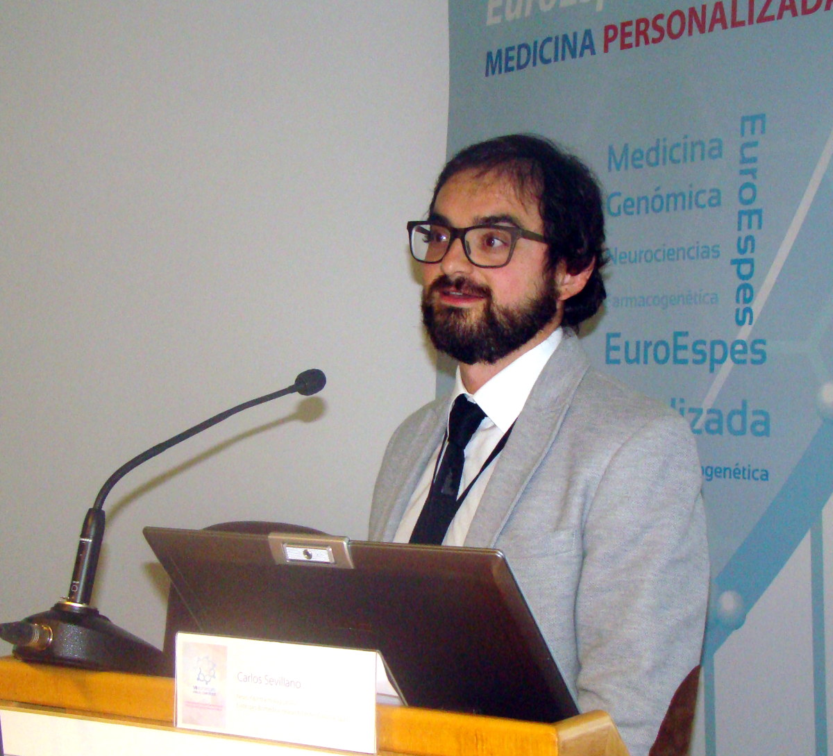Un hombre con barba y gafas habla en un podio con micrófono. Lleva traje gris y corbata negra. Detrás de él hay una pancarta con un texto relacionado con la medicina personalizada y la genómica.
