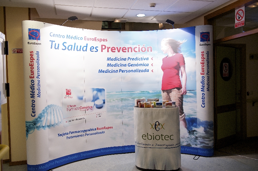 Expositor promocional del Centro Médico EuroEspes que destaca la medicina personalizada y la prevención. En el frente hay una mesa redonda con el logo de "ebiotec" rodeada de folletos. Como fondo se muestran temas relacionados con la salud y una mujer con un vestido blanco.