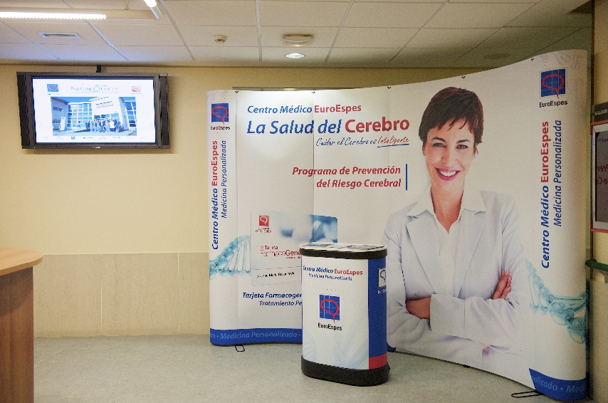 Exposición de un congreso médico en el que aparece una persona sonriente con bata blanca sobre un fondo curvo. El texto incluye "Centro Médico EuroEspes" y "La Salud del Cerebro". También hay un pequeño podio y una pantalla montada en la pared que muestra la imagen de un edificio.