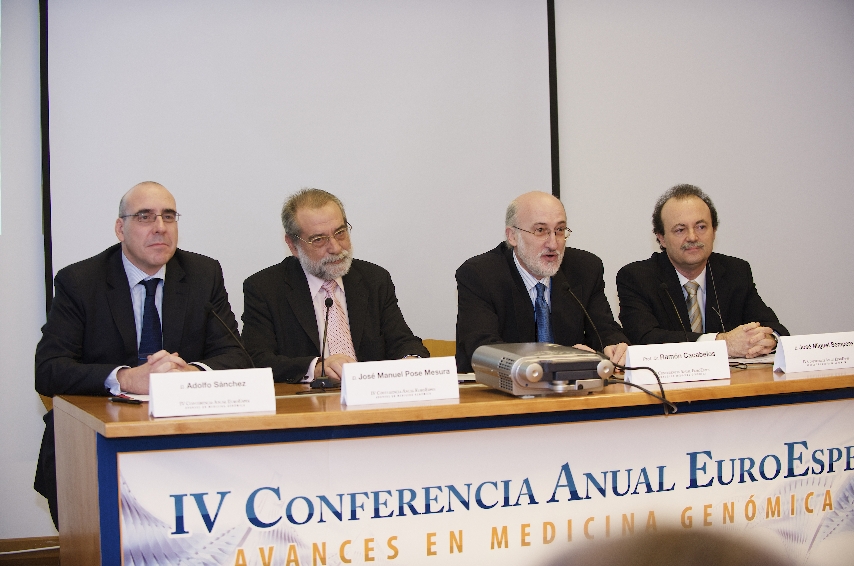 Cuatro hombres de traje se sientan en una mesa de conferencias con micrófonos y placas con nombres. Detrás de ellos hay una pancarta que dice "IV Conferencia Anual EuroEspañola" relacionada con los avances en medicina genómica.