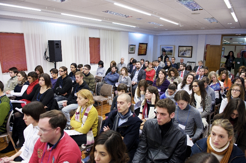 Un grupo grande de personas sentadas en una sala bien iluminada, asistiendo a una conferencia o taller. Algunas personas toman notas, mientras que otras escuchan atentamente. La sala tiene paredes blancas y cuadros enmarcados.