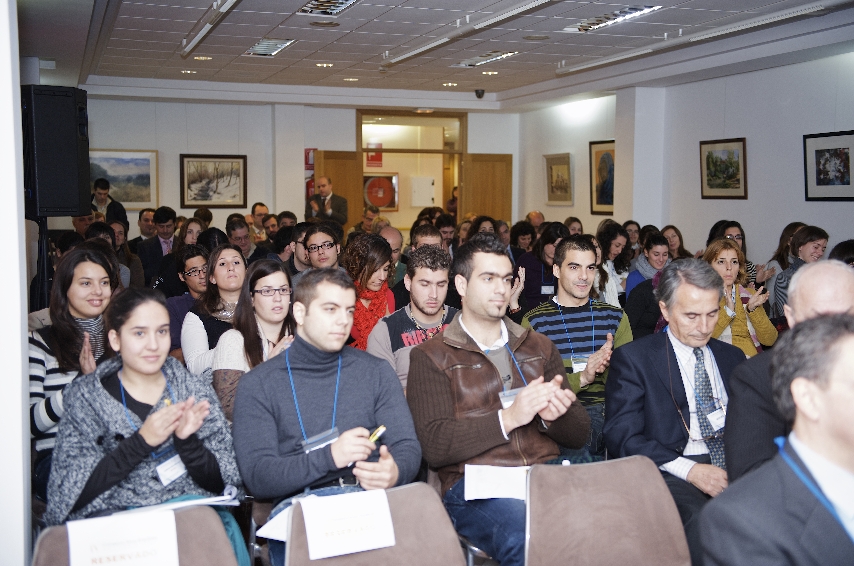 Un gran grupo de personas sentadas en una sala de conferencias, mirando hacia delante. Algunas aplauden, mientras que otras escuchan atentamente. Son de edades y géneros variados, visten ropa informal y de negocios, y llevan etiquetas con sus nombres a la vista. Filas de cuadros cubren las paredes.