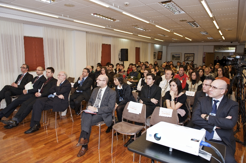 Un grupo de personas vestidas de negocios se sientan atentamente en filas de sillas en una sala de conferencias. En primer plano se ve un proyector. La sala está bien iluminada con suelo de madera y cortinas de colores neutros.