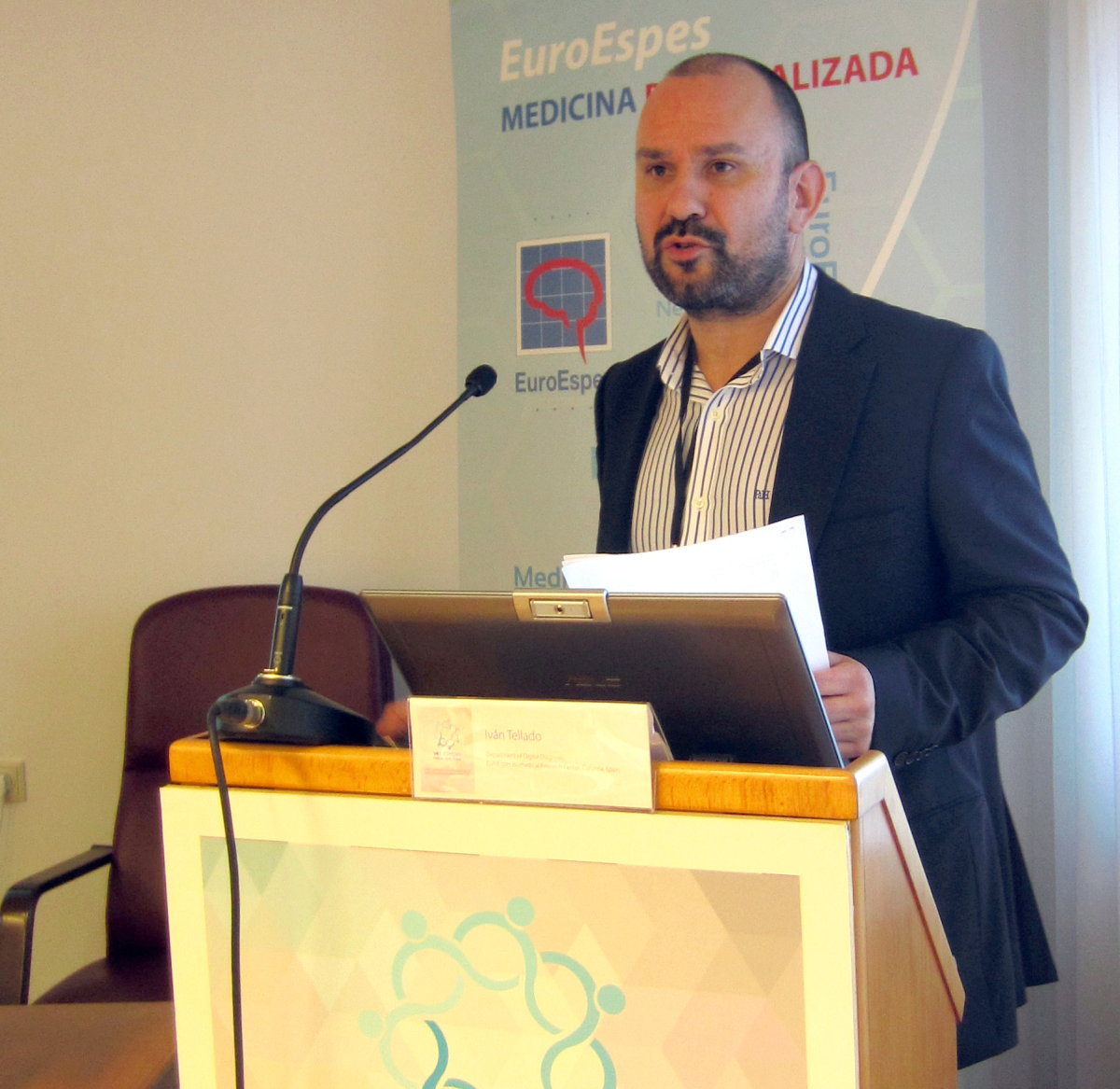 Un hombre de traje habla en un podio, con papeles en la mano. En el fondo se puede leer "EuroEspes Medicina Personalizada", lo que sugiere una conferencia o presentación sobre medicina personalizada.