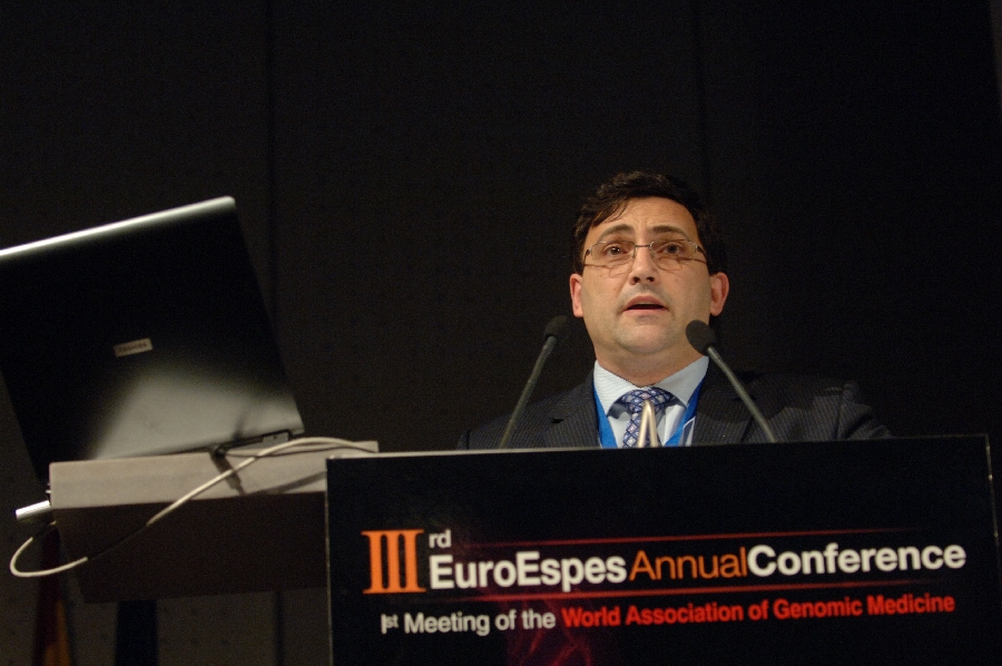 Un hombre con traje y corbata habla en un podio durante la 3.ª Conferencia Anual de EuroEspes. A su lado hay un ordenador portátil abierto. El evento es la 1.ª Reunión de la Asociación Mundial de Medicina Genómica.