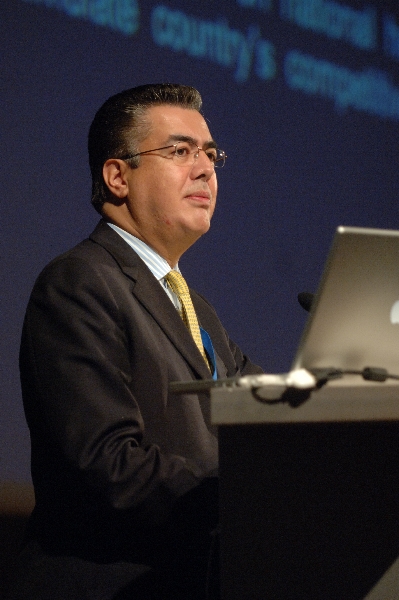 Hombre con gafas y traje hablando en un podio con una computadora portátil, sobre un fondo oscuro.