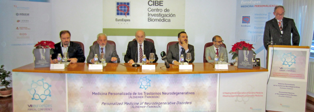Un panel de seis personas se sienta en una mesa larga durante una conferencia. Detrás de ellas hay una pancarta que anuncia la Conferencia Anual sobre Medicina Personalizada para Trastornos Neurodegenerativos. En la sala se ven una pantalla y dos arreglos florales.