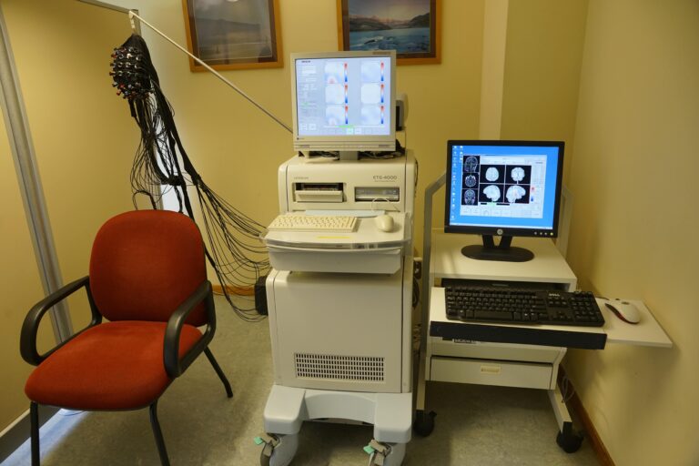 Una silla roja se encuentra junto a una máquina de diagnóstico digital con sensores y cables. Dos monitores muestran imágenes de escáneres cerebrales, lo que refleja las capacidades avanzadas de diagnóstico digital. Cerca de allí, un teclado y un mouse descansan sobre un escritorio en el entorno clínico.