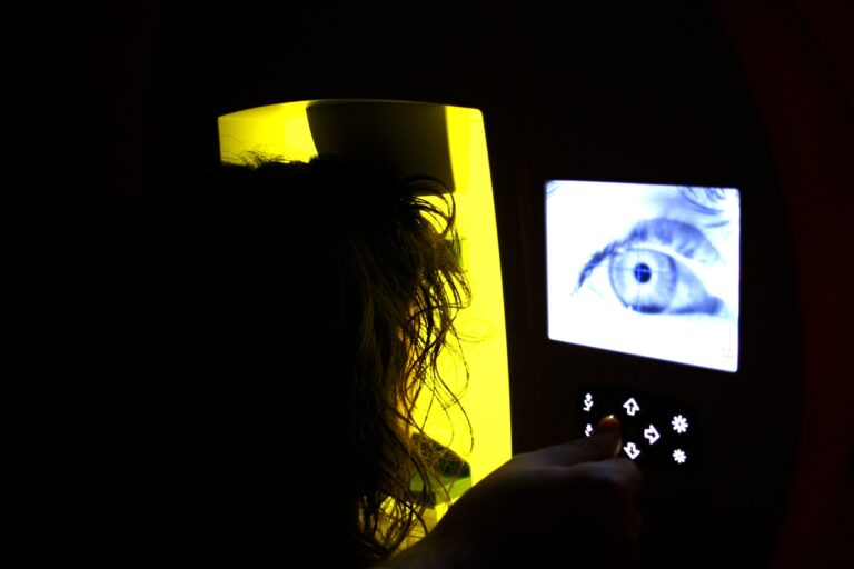 Una persona interactúa con un dispositivo que muestra un gran ojo en la pantalla, que recuerda a la investigación neurooftalmológica. Está en la sombra, parcialmente iluminado por el resplandor amarillo de la máquina. Se ven varios botones táctiles debajo de la pantalla.