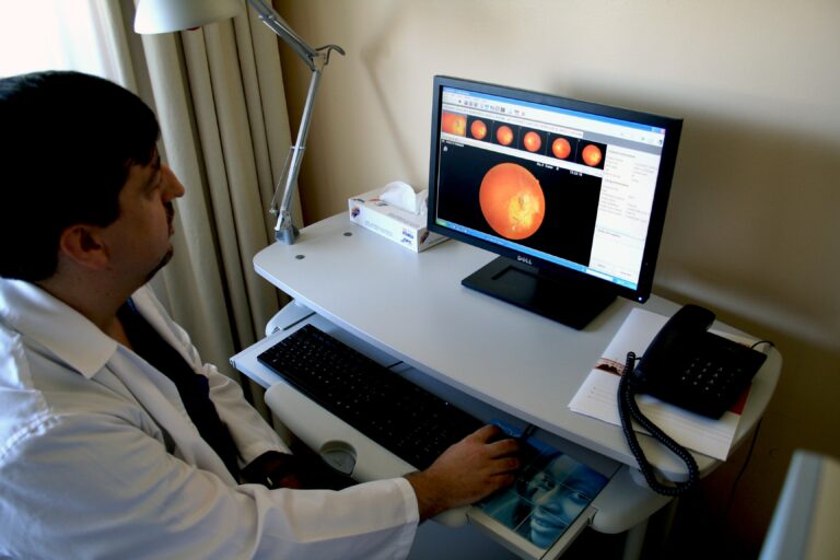 Un médico especialista en neurooftalmología se sienta en un escritorio y examina atentamente un escáner ocular en el monitor de la computadora. La imagen detallada de la retina capta su atención, mientras que un teléfono y una caja de pañuelos descansan cerca, lo que subraya la combinación de tecnología y atención al paciente.