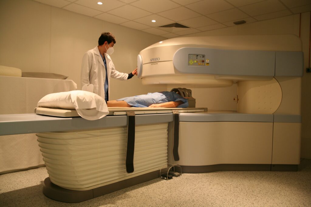 Un paciente se recuesta en una mesa, listo para que le hagan una resonancia magnética en un centro médico. Un profesional sanitario con bata blanca se encuentra de pie junto a la máquina de resonancia magnética, comprobando los controles. La sala está bien iluminada y equipada para realizar procedimientos de diagnóstico por imagen.