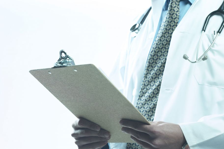 Un médico con bata blanca está de pie con un estetoscopio alrededor del cuello y sujetando un portapapeles. El centro de atención se centra en la parte media del cuerpo, donde se muestran notas sobre enfermedades cerebrovasculares y parte del atuendo del médico, incluida una corbata con un patrón geométrico.