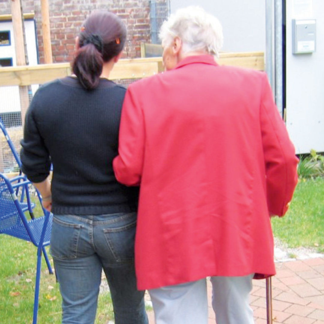 Una mujer con un suéter negro y jeans está de pie junto a una persona mayor con una chaqueta roja que usa un bastón. Están al aire libre en un patio, con césped y un columpio cerca, lo que simboliza la esperanza de un cambio a medida que cambia el paradigma en el tratamiento del Alzheimer.