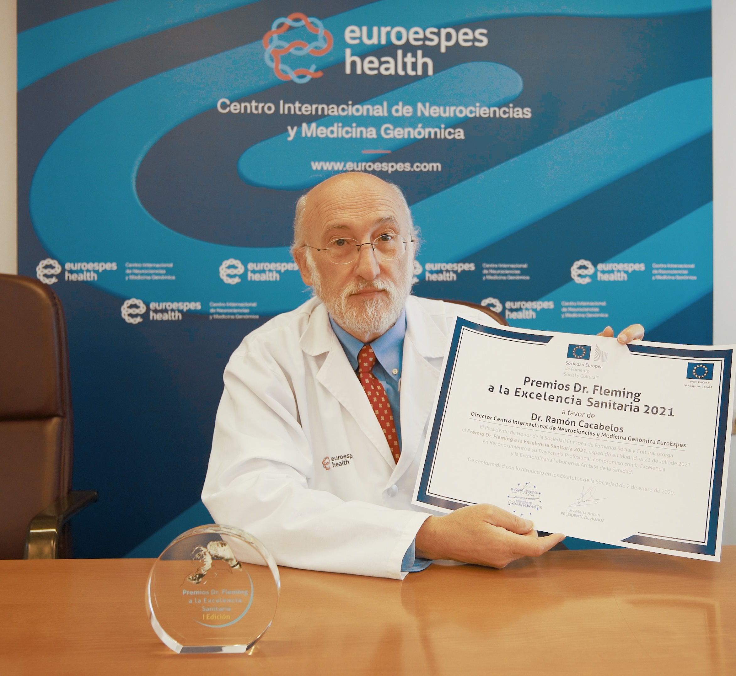 Un hombre con barba y bata blanca sostiene un certificado del "Premio Dr. Fleming a la Excelencia Sanitaria 2021". Está sentado en un escritorio con un premio de cristal, frente a un telón de fondo con una etiqueta que dice "euroespes health".