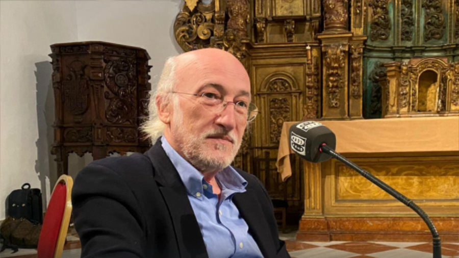 Un hombre mayor con gafas y barba se sienta delante de un micrófono. Lleva una chaqueta de traje oscura y una camisa azul claro. Detrás de él hay una pared decorada con ricos detalles y tonos cálidos. Parece estar enfrascado en una conversación.