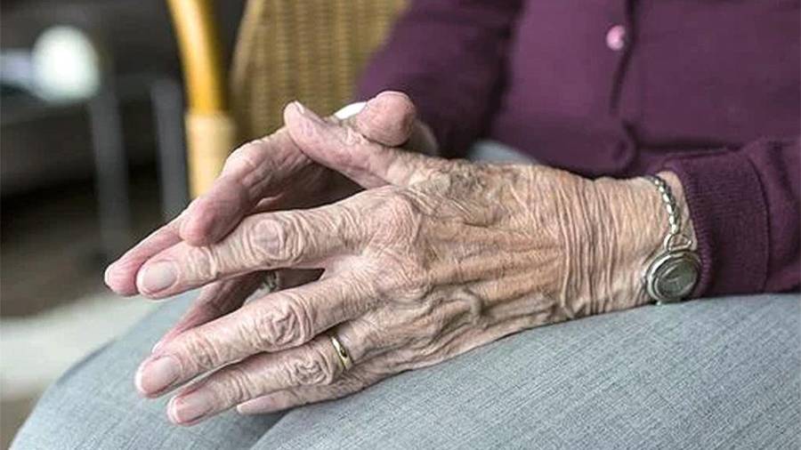 Un primer plano capta las manos entrelazadas de un anciano que descansan sobre su regazo, sutilmente arrugadas y adornadas con un anillo y un reloj. Vestido con un suéter morado y pantalones grises, está sentado tranquilamente en medio de un fondo suavemente borroso, aparentemente intacto por la demagogia emocional con los que estorban.