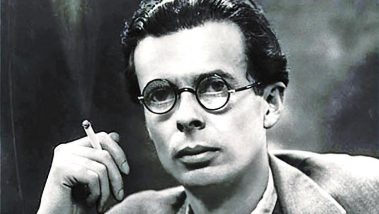 Fotografía en blanco y negro de una persona que lleva gafas redondas y sostiene un cigarrillo cerca de la cara. El individuo mira directamente a la cámara con expresión seria. Está vestido con una chaqueta y una camisa.