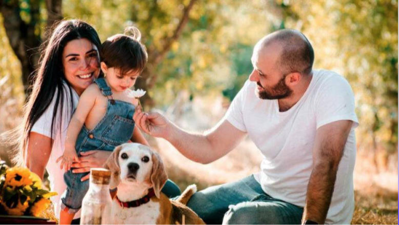 Un hombre y una mujer sonrientes se sientan al aire libre con un niño pequeño y un perro. El hombre sostiene una flor cerca de la nariz del niño. La luz del sol se filtra a través de los árboles del fondo, creando una atmósfera cálida y alegre.