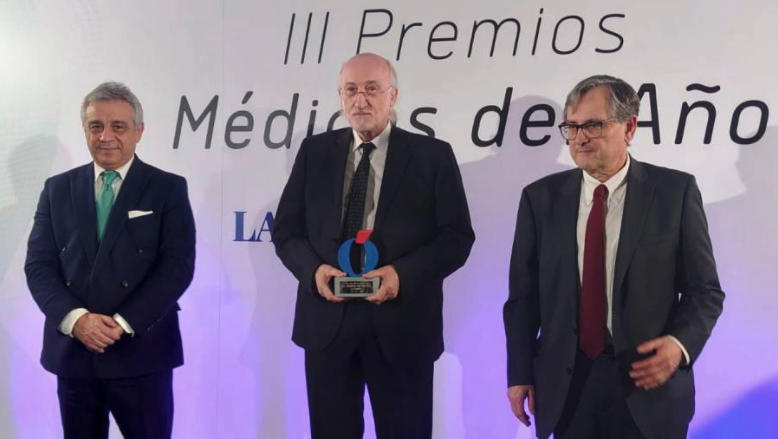 Tres hombres de traje se encuentran de pie frente a un telón con el texto "III Premios Médicos del Año". El hombre del medio sostiene un trofeo. Parecen estar en una ceremonia de entrega de premios.
