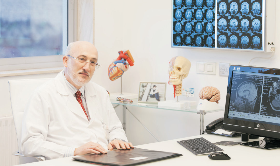 Un médico con bata blanca se sienta en un escritorio en un consultorio médico, personificando el enfoque de Euroespes marca el rumbo hacia la medicina personalizada. Detrás de él hay imágenes médicas, modelos anatómicos del corazón y el cerebro y una computadora que muestra imágenes cerebrales. La luz natural se filtra a través de la ventana de la izquierda.