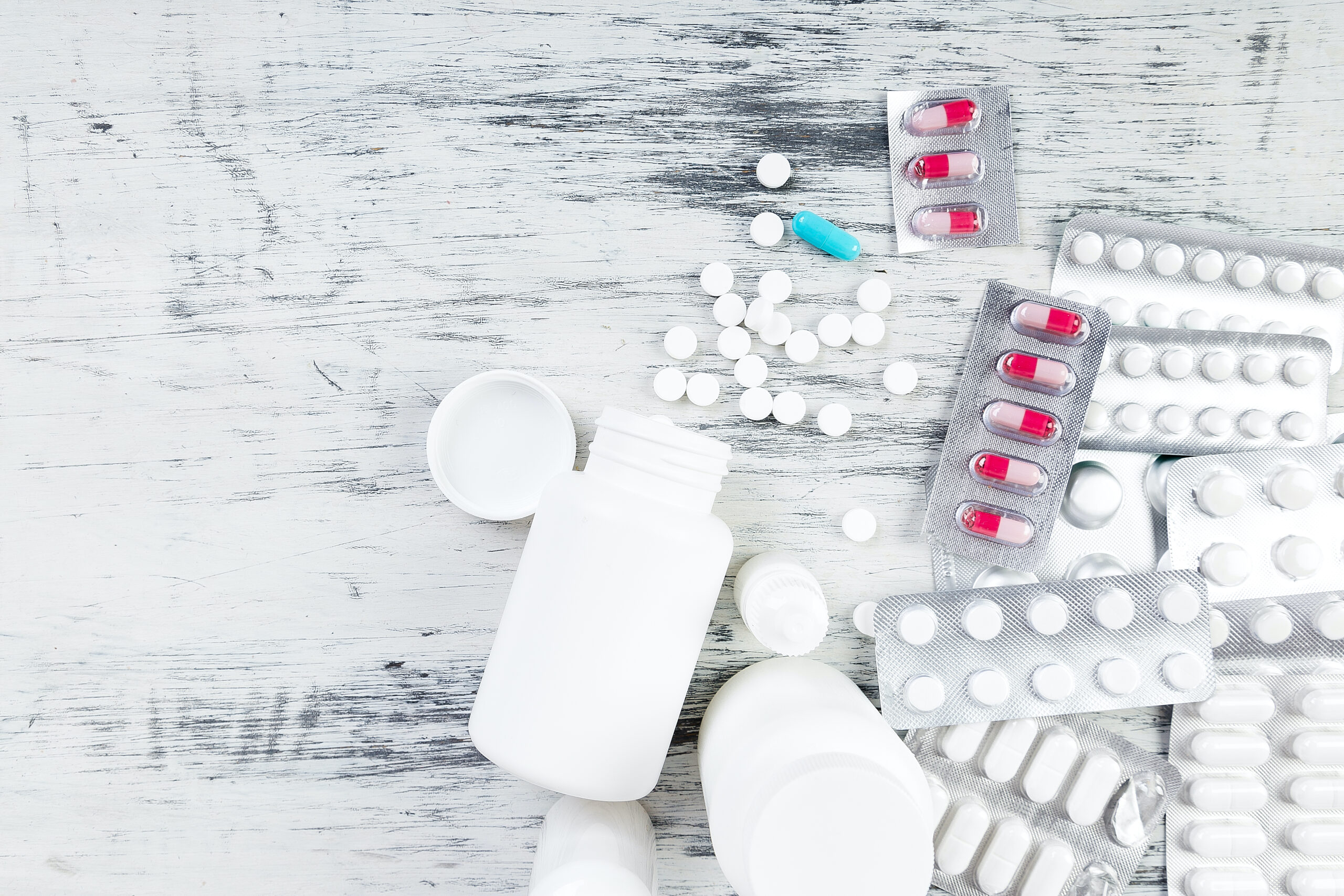 Una selección de diversos medicamentos, entre ellos pastillas, comprimidos y cápsulas, en blísters y en envases sueltos sobre una superficie de madera blanca envejecida. Algunos frascos de plástico blancos están abiertos y vacíos, lo que aumenta el surtido.