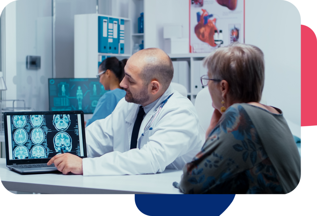 En un consultorio médico moderno, un médico muestra imágenes cerebrales en una computadora portátil a un paciente, destacando los servicios médicos avanzados. En el fondo, otro profesional de la salud trabaja diligentemente en una computadora, asegurando una coordinación perfecta.