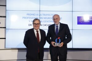 Dos hombres de traje se encuentran frente a una pantalla que muestra un texto en español sobre un premio a la trayectoria profesional. Uno de ellos sostiene un trofeo mientras los dos sonríen para la foto.