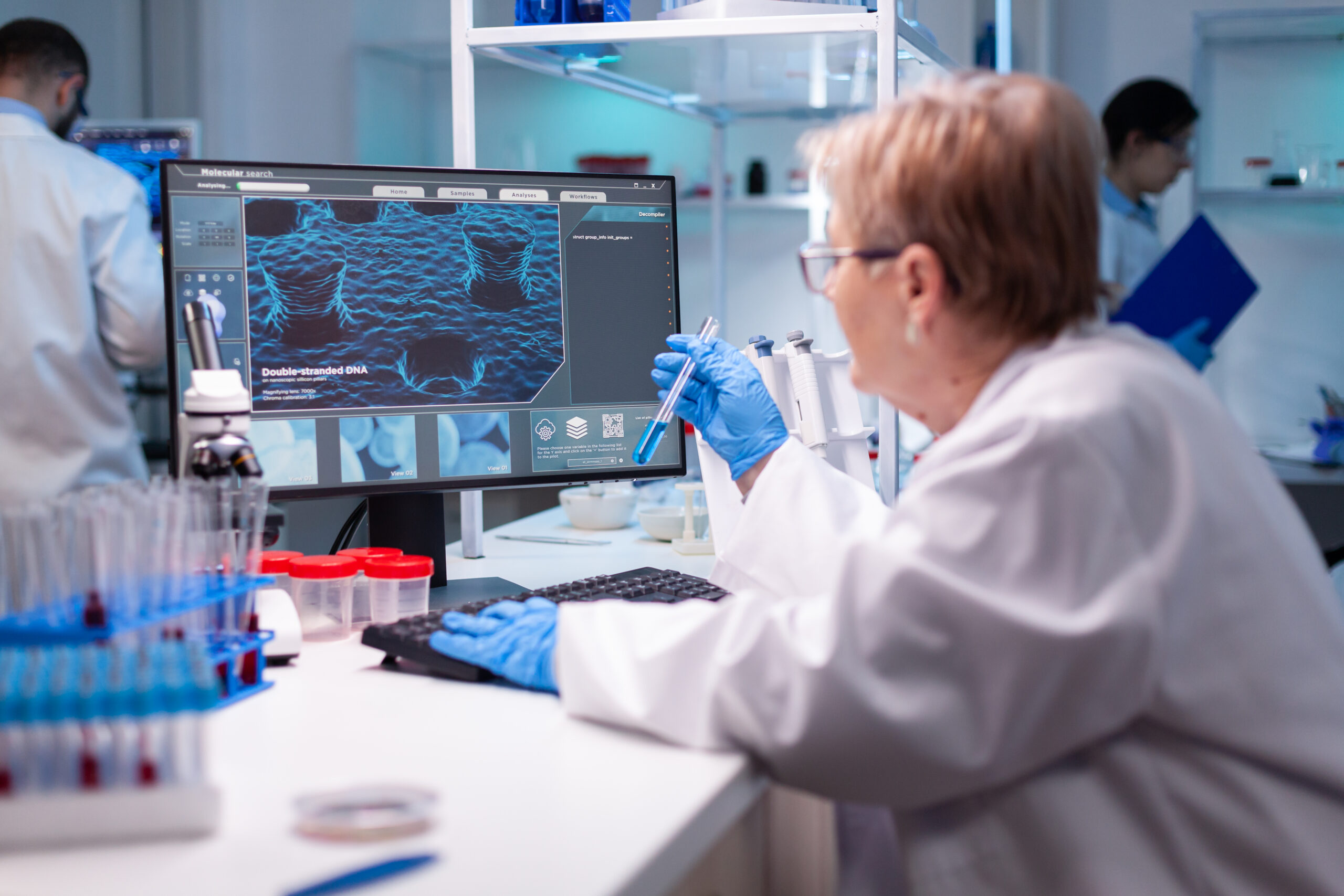 Un científico con bata y guantes está sentado en un escritorio analizando las estructuras de ADN que se muestran en un monitor de computadora. El laboratorio está lleno de equipos y tubos de ensayo, lo que sugiere un entorno de investigación centrado en estudios genéticos.