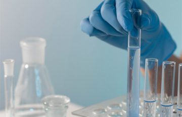 Una mano enguantada sostiene un tubo de ensayo que contiene un líquido transparente sobre una gradilla llena de más tubos de ensayo, encarnando la precisión de la que se habla a menudo en el Boletín Médico EuroEspes. Al fondo, se ven diversos equipos de laboratorio de vidrio sobre una superficie suavemente iluminada, evocando una atmósfera de salud e innovación científica.