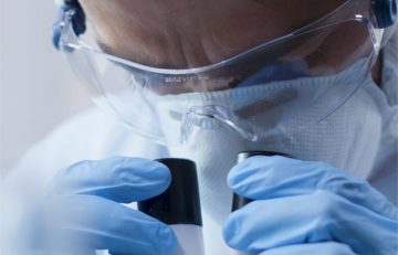 Una persona con gafas protectoras, mascarilla y guantes azules mira diligentemente a través de un microscopio, lo que resalta el compromiso con la seguridad y la precisión en este entorno de laboratorio de salud del Boletín Médico EuroEspes.