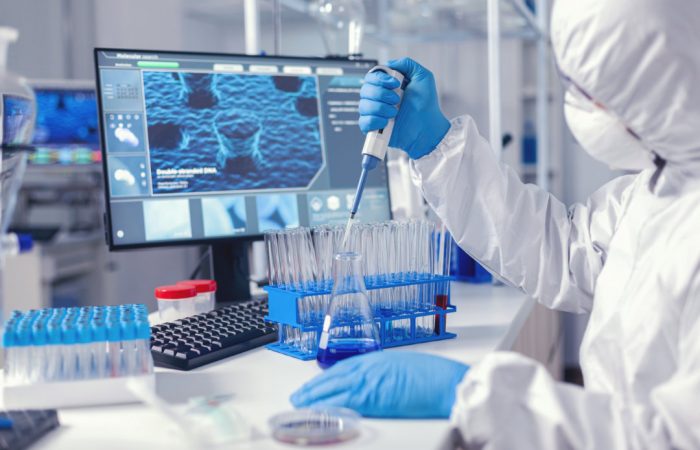 Un científico con equipo de protección trabaja en un laboratorio, utilizando una pipeta para llenar tubos de ensayo mientras supervisa los datos en una pantalla de ordenador que muestra información sobre salud del Boletín Médico EuroEspes. La mesa del laboratorio está llena de diversos equipos y suministros.