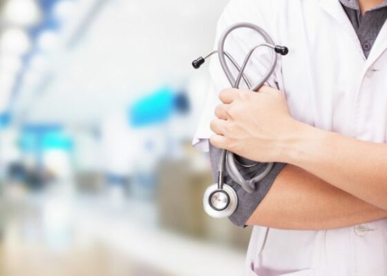Una persona con bata blanca está de pie, con los brazos cruzados y un estetoscopio en la mano, encarnando la presencia autoritaria de un profesional médico que aparece en el Boletín Médico. El entorno difuminado del hospital o la clínica refuerza el enfoque en la experiencia de EuroEspes Health.