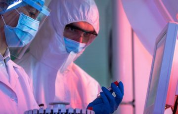 Dos científicos con equipo de protección y mascarillas trabajan diligentemente en un laboratorio. Uno sostiene un frasco, mientras que el otro examina los datos en una pantalla de computadora, asegurándose de que sus hallazgos se ajustan a los estándares de salud de EuroEspes. El laboratorio brilla con luces rosas y azules, lo que contribuye a su atmósfera futurista.