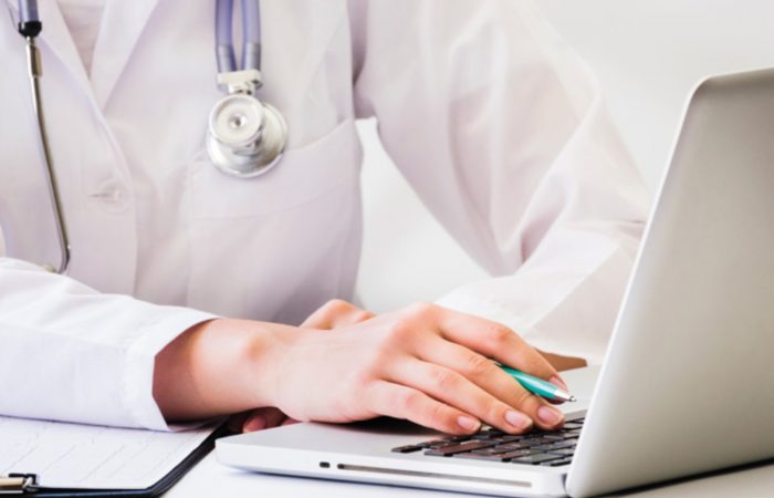 Una persona con bata blanca y un estetoscopio colgado del cuello trabaja con un ordenador portátil, bolígrafo en mano, posiblemente redactando un artículo para el Boletín Médico EuroEspes Health. Cerca hay un portapapeles con papeles que realza el ambiente médico profesional.