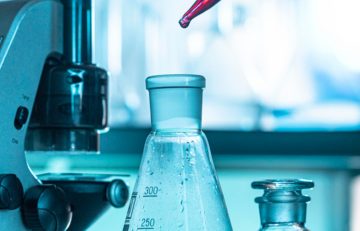 Primer plano de un equipo de laboratorio, incluido un microscopio y material de vidrio, que aparece en el Boletín Médico EuroEspes. Un gotero libera un líquido rojo en un matraz, con otro recipiente cerca, todo ello en un entorno de laboratorio científico que muestra investigaciones sanitarias de vanguardia.
