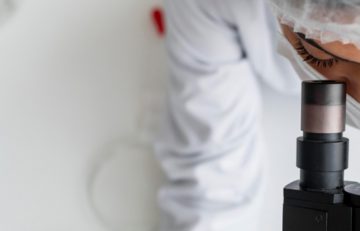 Un científico vestido con una bata blanca y una redecilla para el pelo observa a través de un microscopio en el laboratorio de Salud de EuroEspes, con intensa atención y concentración en la tarea en cuestión.