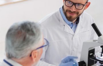 En el luminoso laboratorio, dos científicos con batas de laboratorio están inmersos en una discusión. Uno, con gafas y barba, está de pie junto a un microscopio; el otro sostiene un tubo de ensayo con las manos enguantadas. Su conversación refleja las ideas del Boletín Médico EuroEspes Health, lo que subraya su compromiso y concentración.