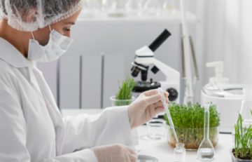 Un científico con bata, mascarilla y redecilla para el pelo realiza un experimento con tubos de ensayo, rodeado de pequeñas plantas y un microscopio sobre una mesa de laboratorio blanca. Esta minuciosa investigación podría aparecer próximamente en el Boletín Médico EuroEspes, lo que contribuirá a mejorar nuestra comprensión de la salud.