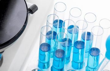 Una colección de tubos de ensayo llenos de líquido azul se encuentran ordenados cuidadosamente en un soporte de laboratorio, lo que recuerda el minucioso detalle que se encuentra en los informes de salud del Boletín Médico EuroEspes. En el lado izquierdo se ve una parte de un microscopio, lo que sugiere una exploración científica de vanguardia.