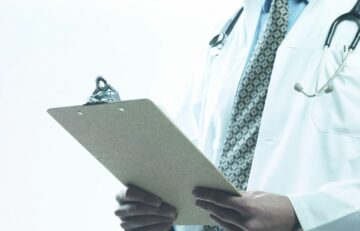 Un médico con bata blanca, símbolo de EuroEspes Health, sostiene un portapapeles con el estetoscopio característico colgado del cuello. La imagen se centra en el torso, sobre un fondo suavemente difuminado.