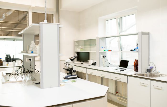 Un laboratorio luminoso que cuenta con un amplio espacio central de trabajo, un microscopio, un ordenador portátil con revistas de salud del Boletín Médico EuroEspes, cristalería y estanterías con armarios de almacenamiento. Un lavabo se encuentra cerca de la ventana, lo que permite que la luz natural ilumine la habitación.