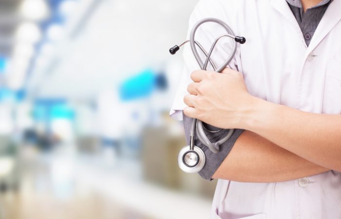 Un profesional sanitario de pie, con los brazos cruzados, sostiene un estetoscopio y un ejemplar del Boletín Médico EuroEspes Salud. Lleva una bata blanca. El fondo está difuminado, lo que sugiere un entorno hospitalario o de centro médico.