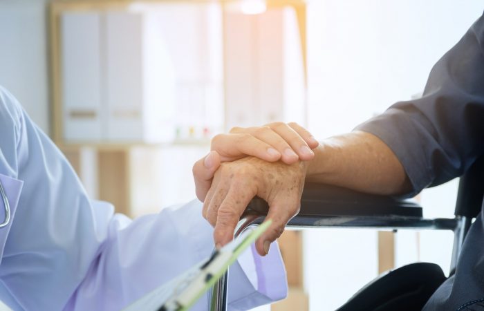 Un profesional sanitario sostiene con delicadeza la mano de un paciente anciano sentado en una silla de ruedas. La escena transmite atención y apoyo, con un fondo luminoso y tenue que sugiere un entorno médico o de rehabilitación.